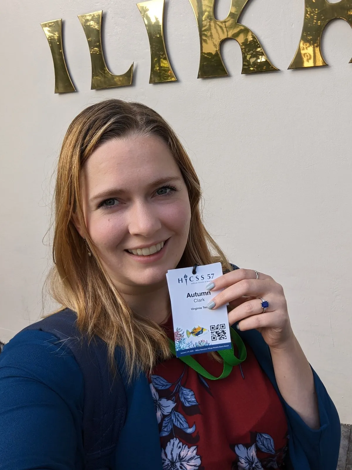 Autumn Clark smiling and holding up her HICSS-57 conference badge outside the conference hotel.
