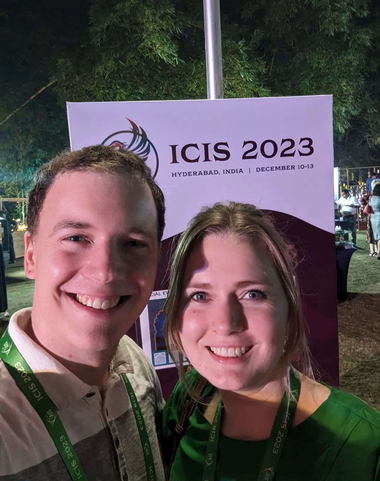 Autumn Clark and another attendee, both wearing ICIS 2023 lanyards, smiling in front of an ICIS 2023 Hyderabad sign at an evening event.
